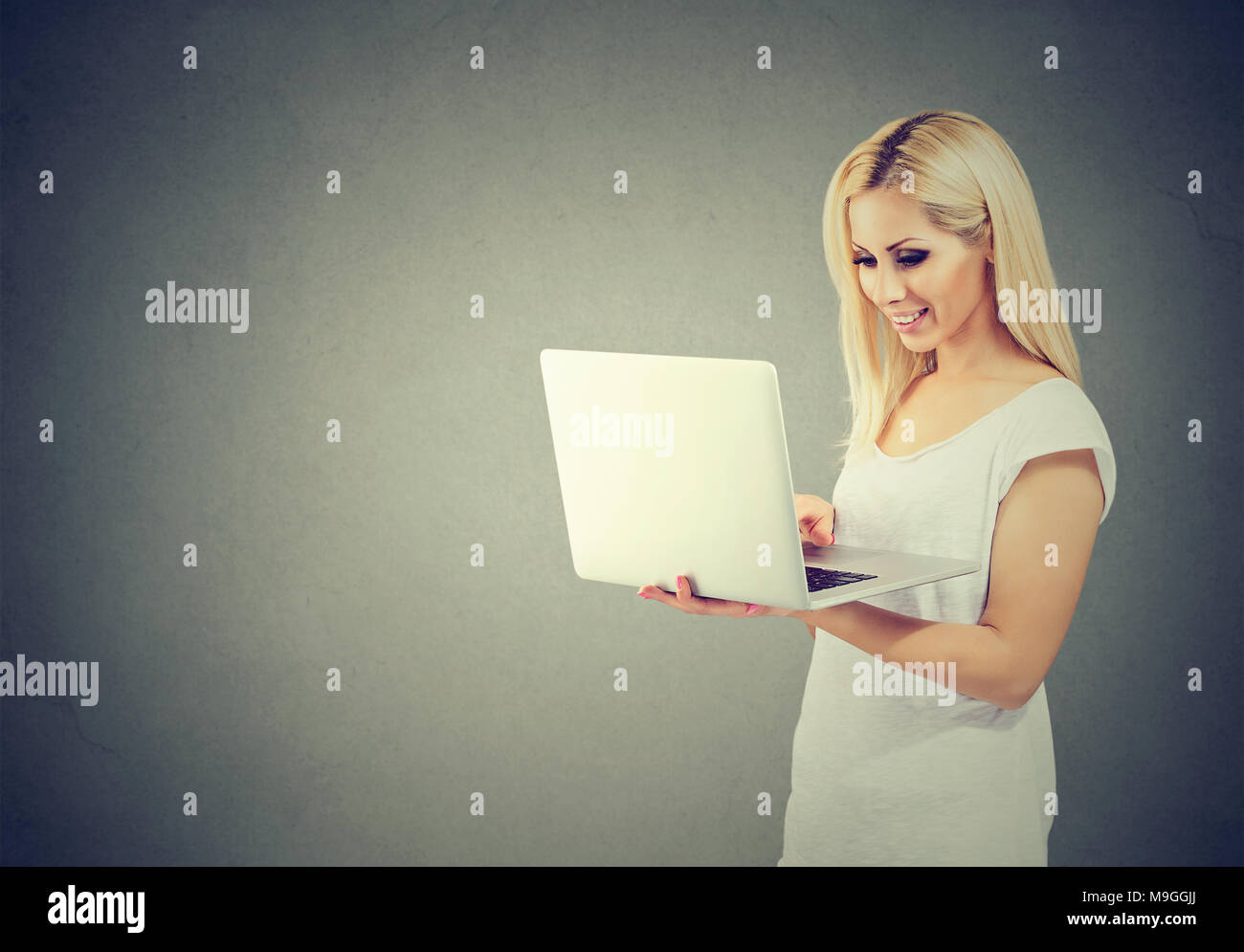 Casual young blonde shopping online using laptop and smiling happily on gray backdrop. Stock Photo