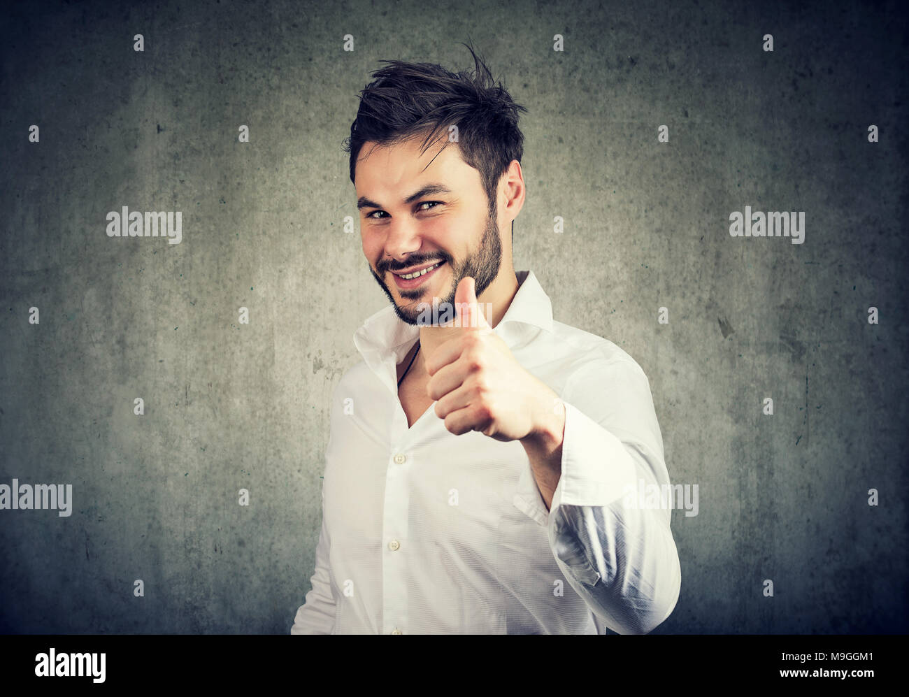 Successful young man in white shirt giving like and showing thumb up with smile. Stock Photo