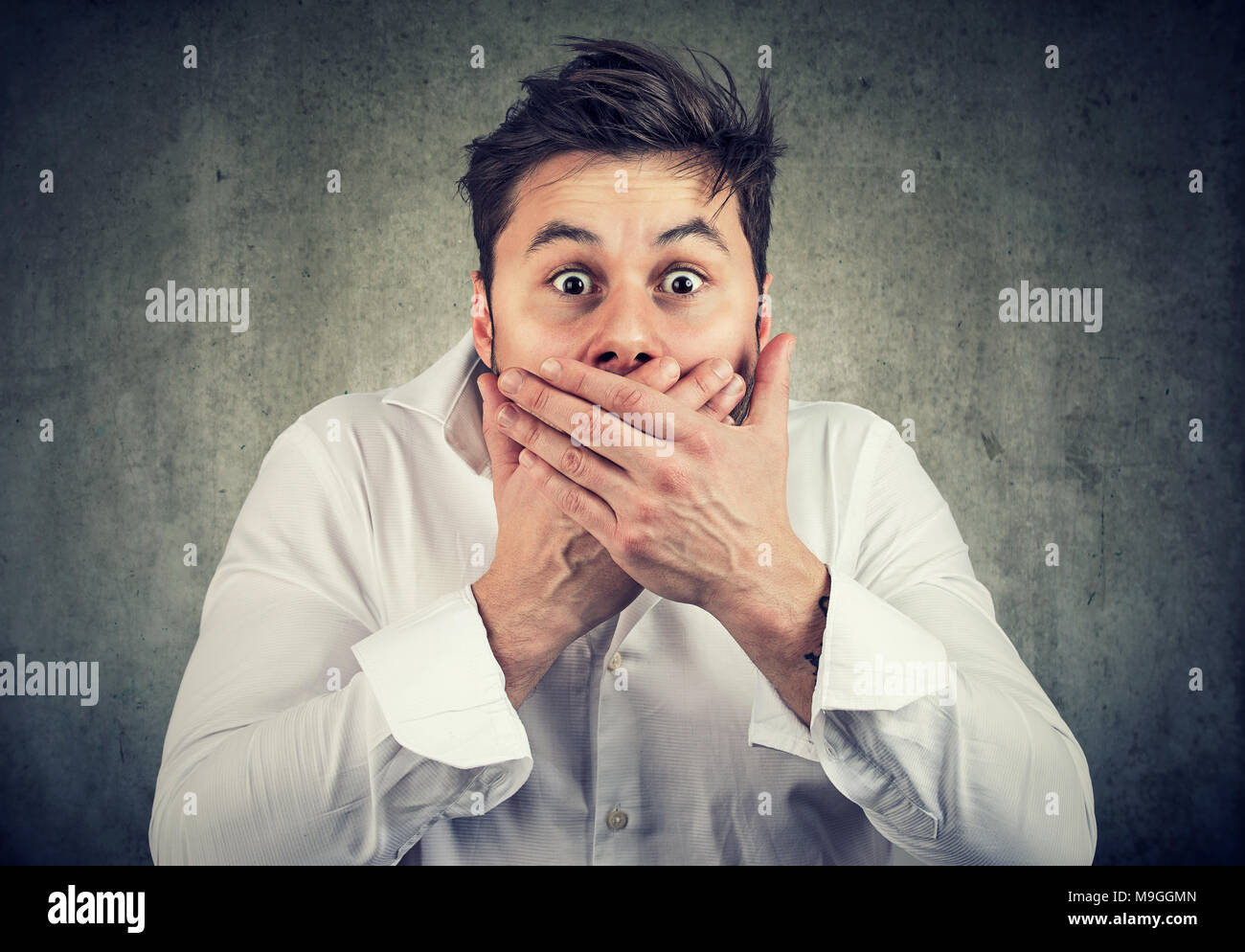 Young shocked man in white shirt covering mouth holding scream while looking at camera with amazed face expression. Stock Photo
