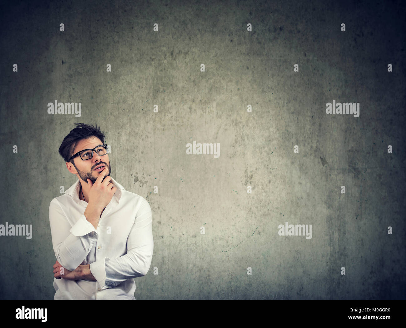 Handsome stylish man in white shirt and eyeglasses touching chin and daydreaming on gray. Stock Photo