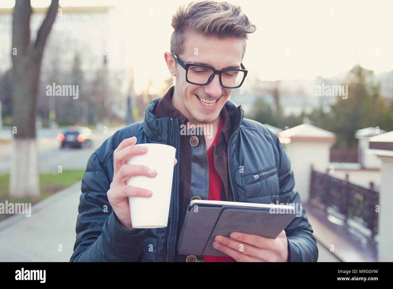 Young smiling man in outwear and eyeglasses using tablet and drinking coffee on street. Stock Photo