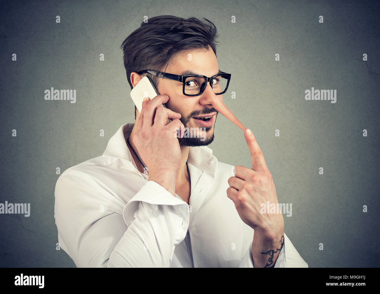 Young handsome man with long nose of liar cheating while speaking on smartphone and looking at camera. Stock Photo