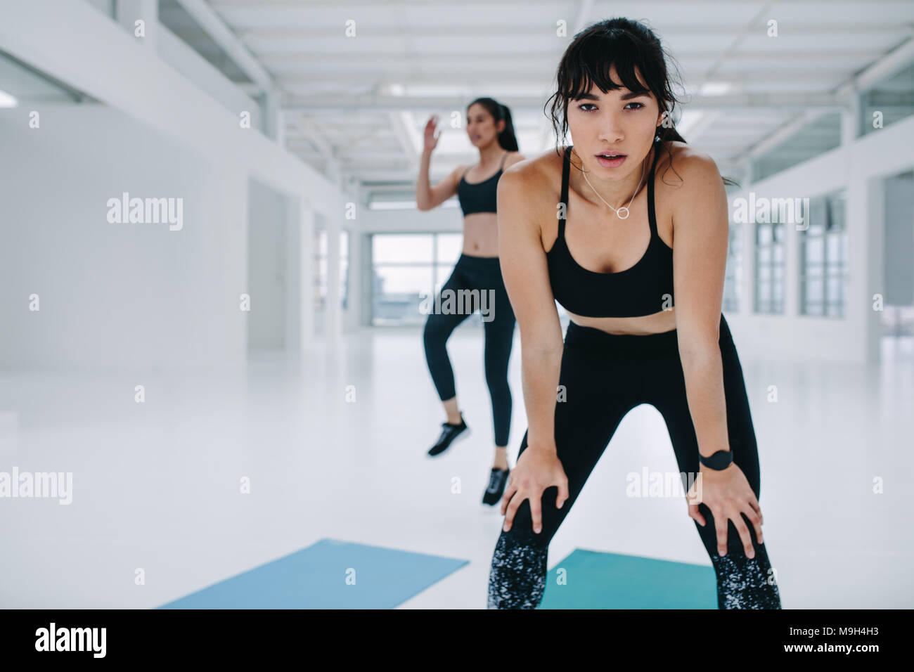 Sportswoman in bent forward pose with hands on knees and female friends exercising in background. Woman taking break in exercise class in gym. Stock Photo