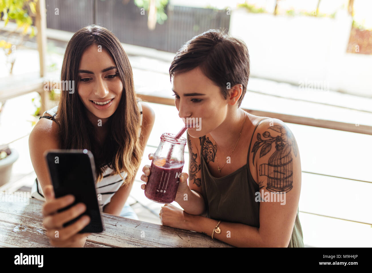 Two women sitting at a restaurant looking at a mobile phone. Woman showing mobile phone while another woman enjoys a smoothie. Stock Photo