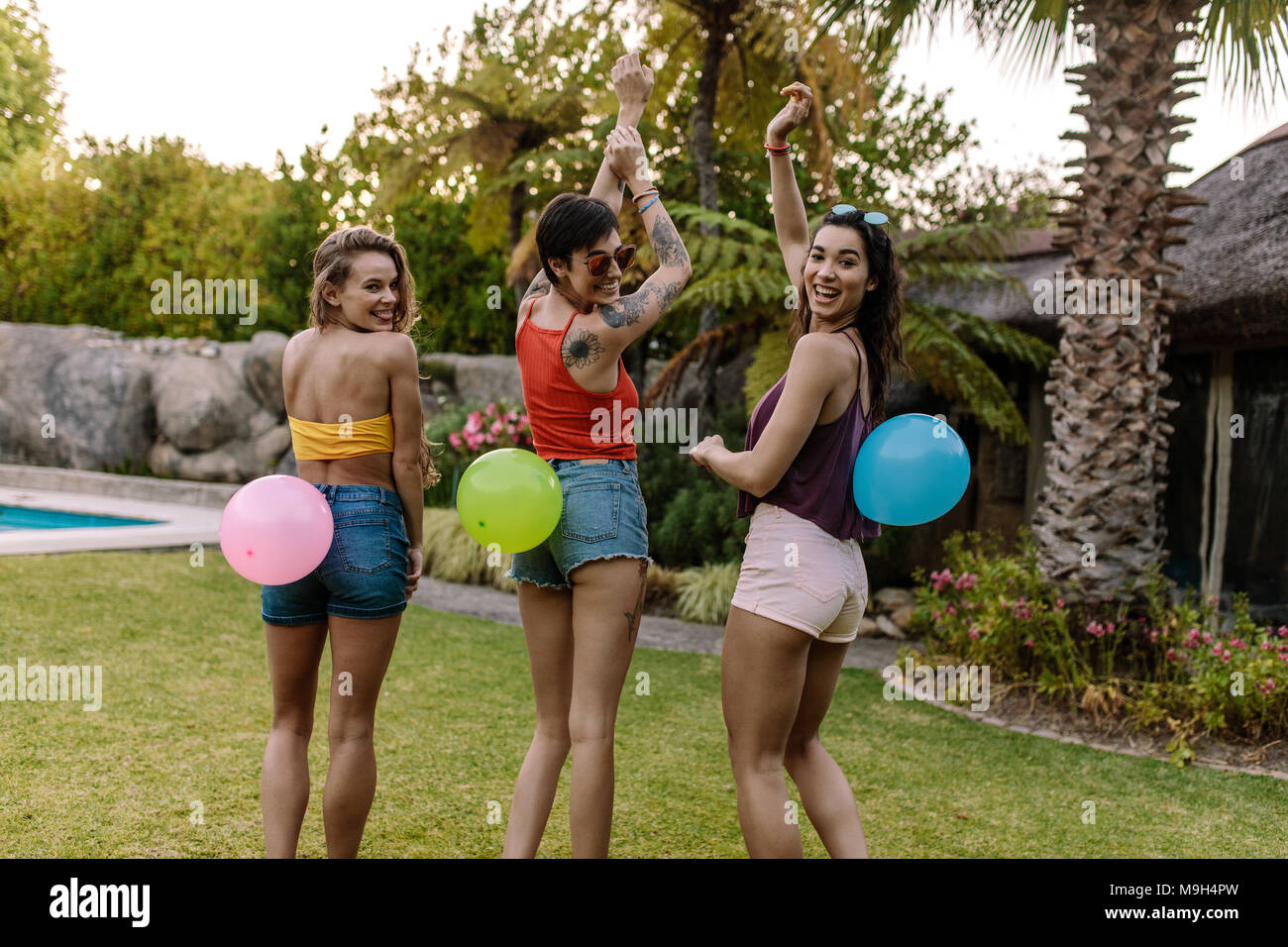 Smiling women standing outdoors with balloons tied on their back. Group of friends enjoying balloon pop game at party. Stock Photo