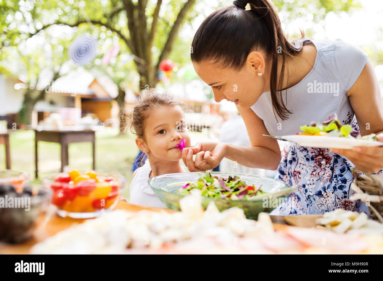 Family celebration outside in the backyard. Big garden party. Birthday party. Young mother with a small girl standing at the table with food. Stock Photo