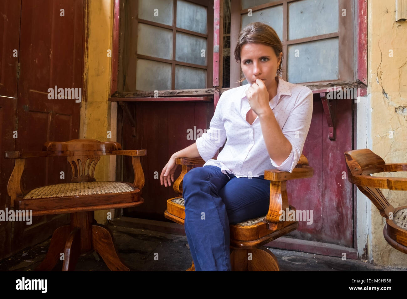 Caucasian young woman sitting in retro armchair in strange old shabby place and looking at camera Stock Photo