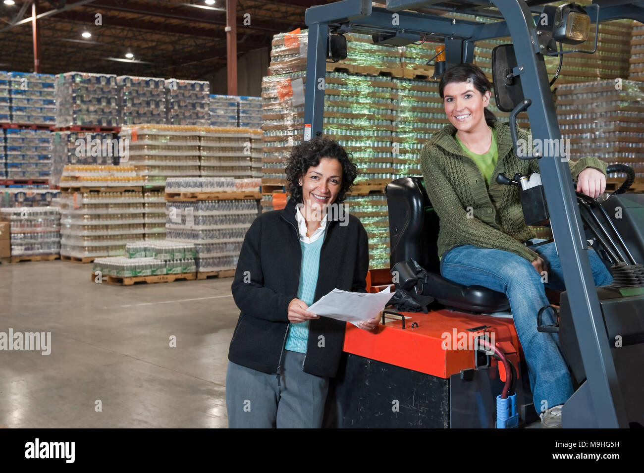 Portrait of two multi-ethnic female warehouse workers on a forklift in a large warehouse full of pallets of flavored water in cans. Stock Photo