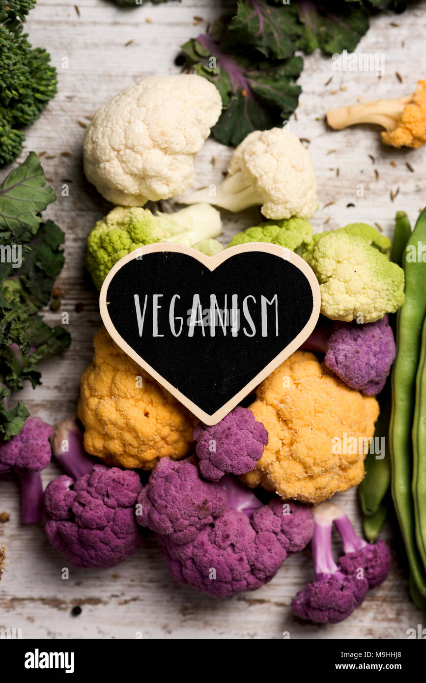 a heart-shaped signboard with the text veganism placed on a pile of some different raw vegetables, such as cauliflower of different colors, broccolini Stock Photo