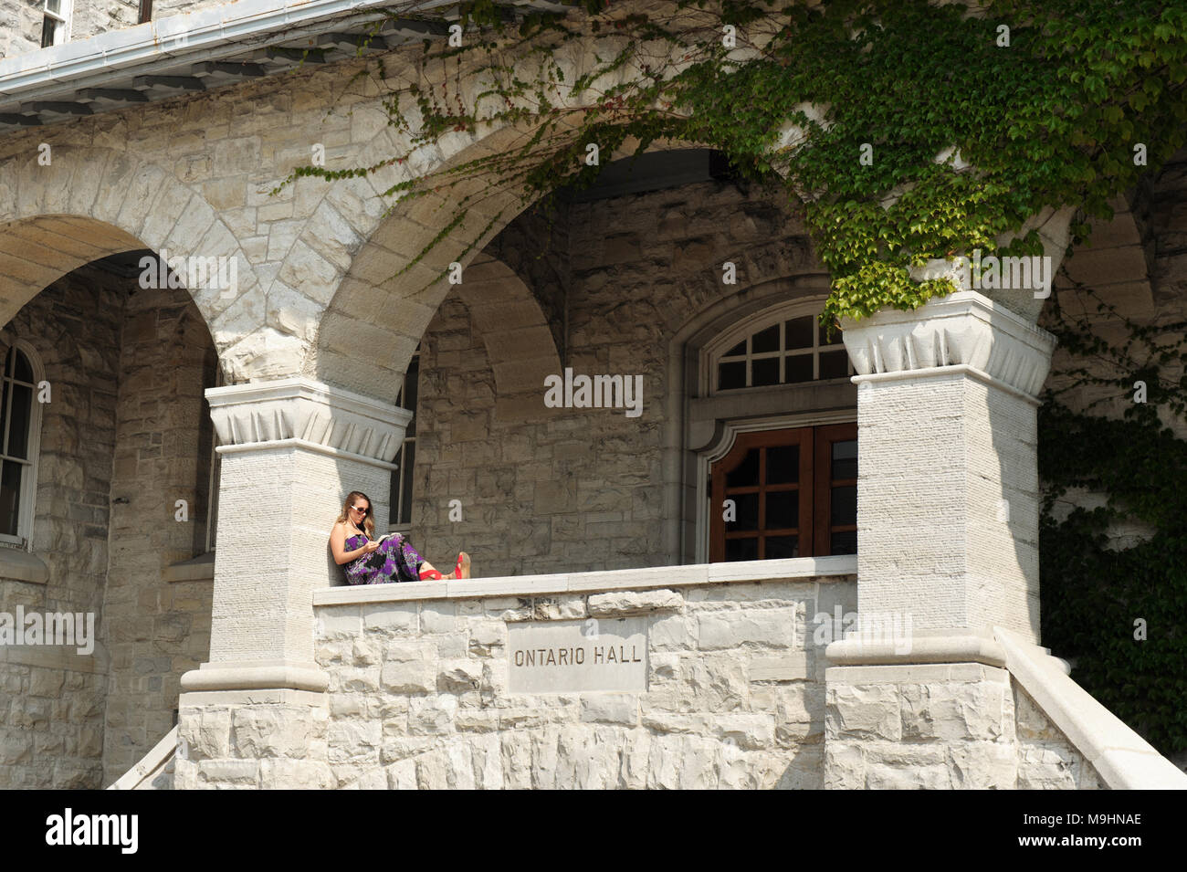 A young woman reads a book, sitting on the porch of Ontario Hall Building, Queen's University, Kingston, Ontario, Canada. Stock Photo