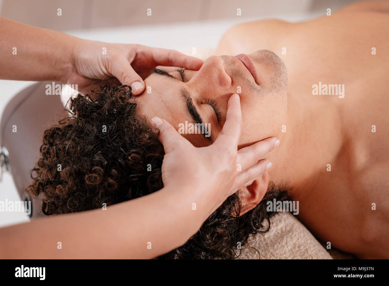 Close-up of a handsome healthy young man enjoying relaxing facial massage at beauty salon. Stock Photo