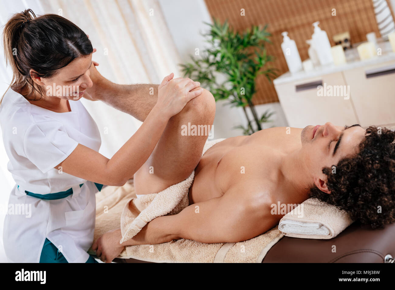 Handsome man lying down on his back, getting healthy leg massage by young female therapist, in spa center. Stock Photo