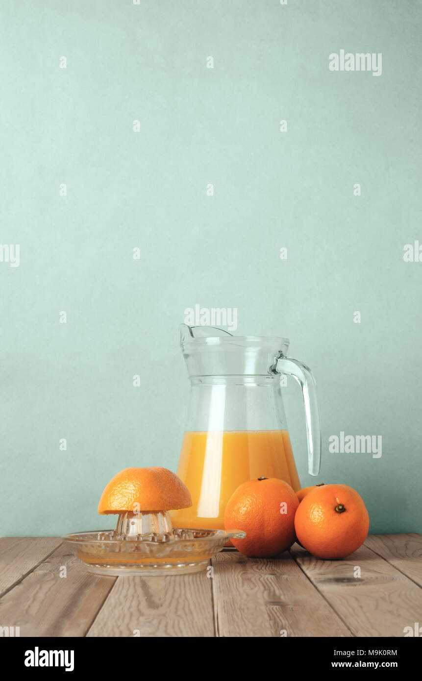 Whole oranges and a half orange on manual squeezer with a glass jug of orange juice behind on a wood plank table with pale turquoise background. Filte Stock Photo