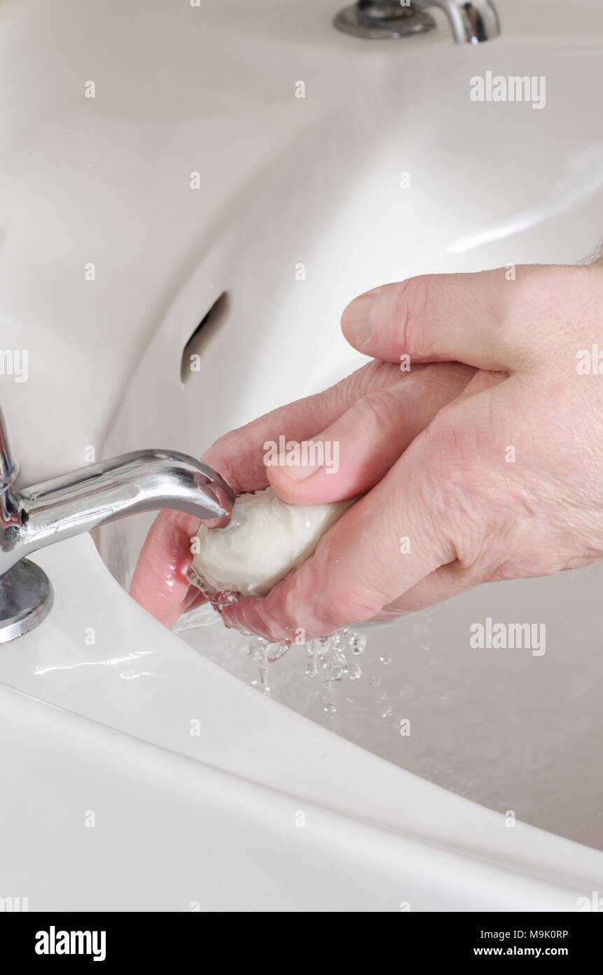 Male hands, holding a white bar of soap and washing under clear running tap water in porcelain sink. Separate hot and cold taps. Stock Photo