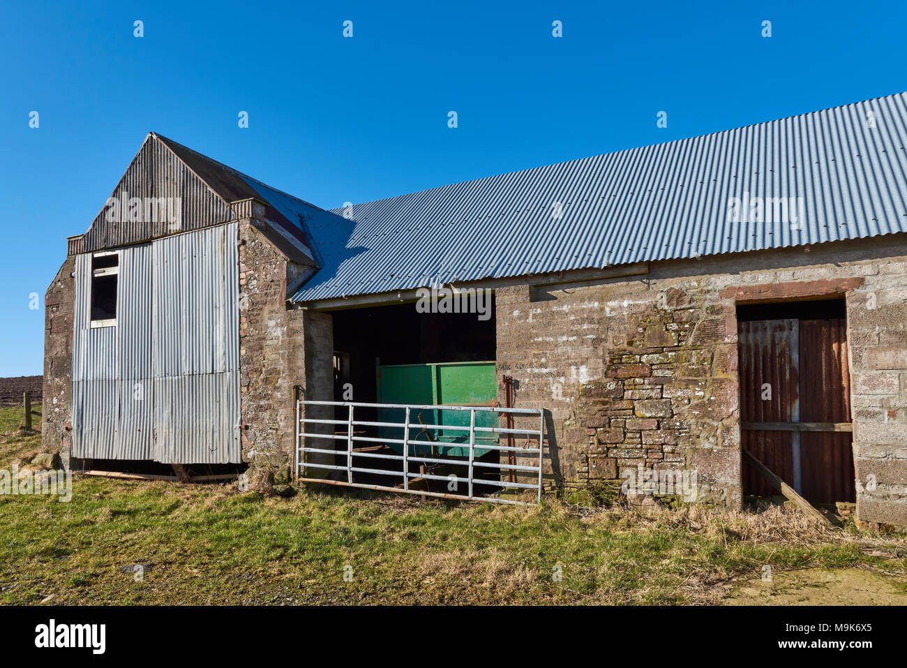 A Green Farm Trailer lies parked within this old Farm Steading, secured only by a simple Farm Gate across a Double Doorway under a corrugated Roof. Stock Photo