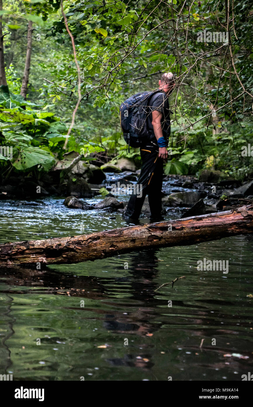 A man with backpack is making his way through the river in a dense forest. Concept of exploration and survival in the wilderness. Stock Photo