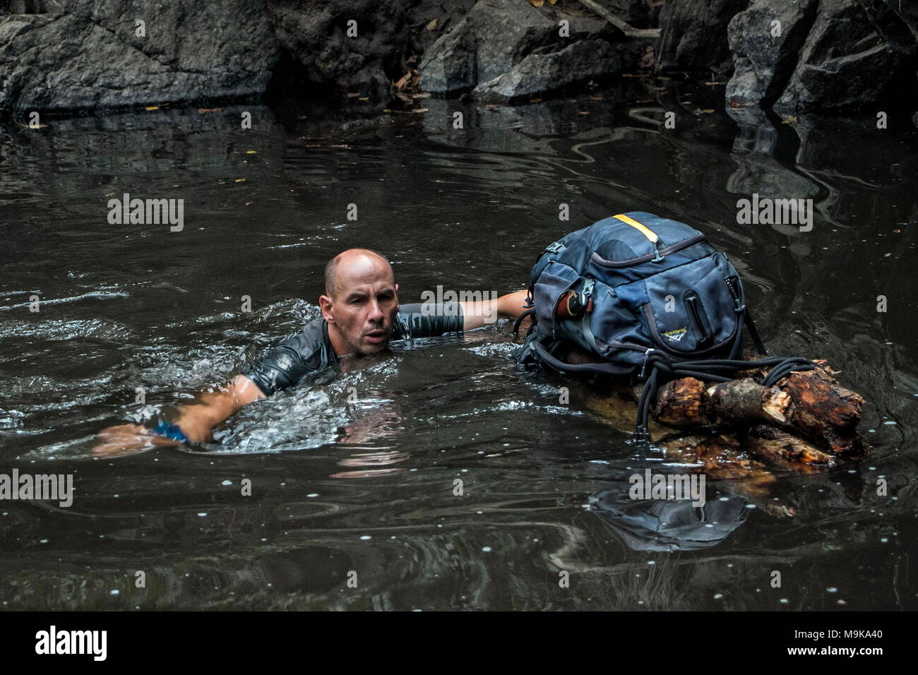 A man is swimming in a river and supporting a hand-made improvised raft with his backpack on. Concept of survival and exploration in the wilderness Stock Photo