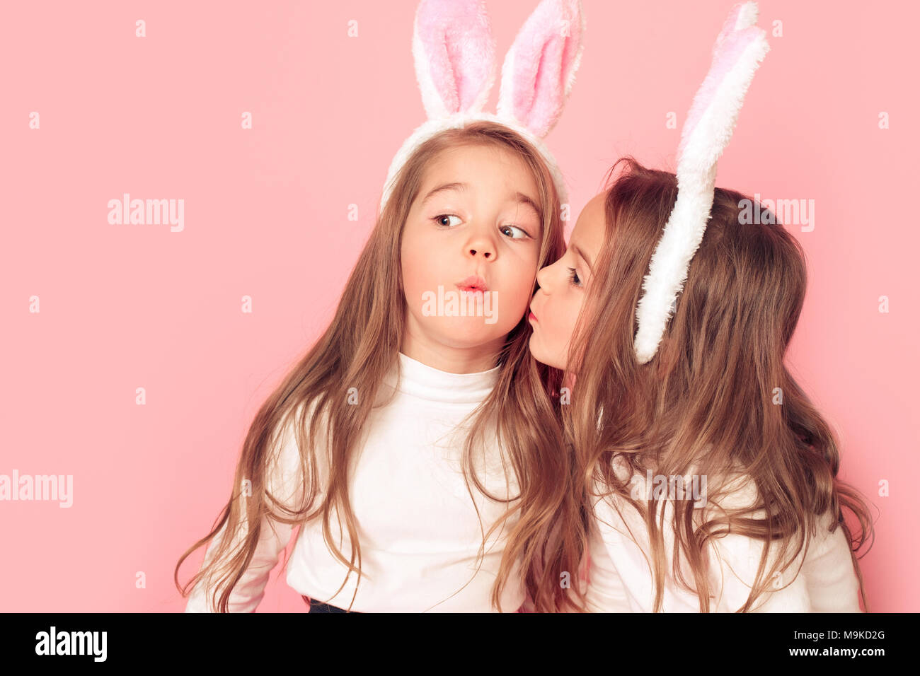 Two little children girls wearing bunny ears studio isolated on pink background easter celebration one girl kissing another cute close-up Stock Photo