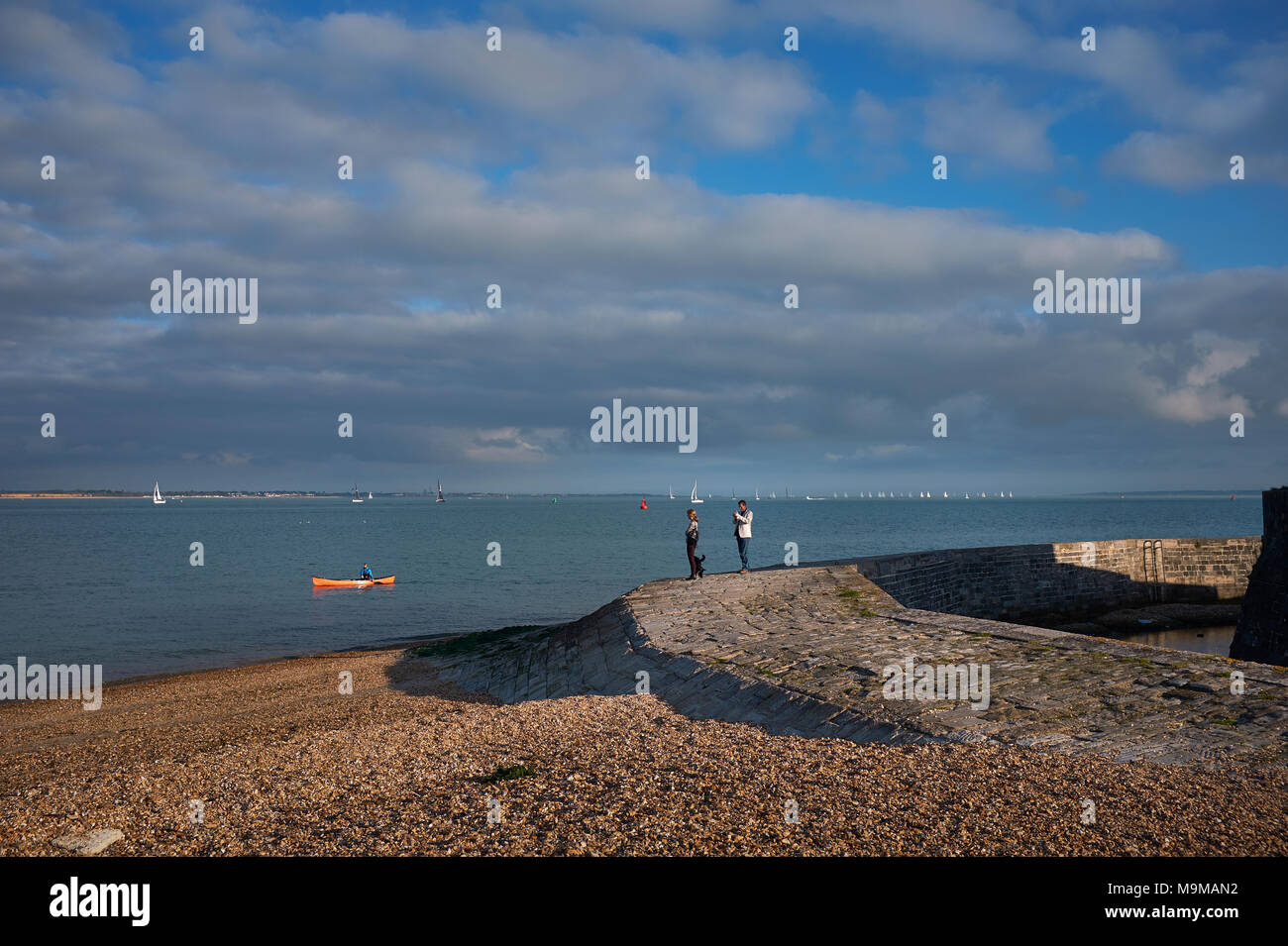 Two people watching a single canoe traveling along the Solent in the English Channel from Calshot beach, UK, with sailing boats in the background Stock Photo