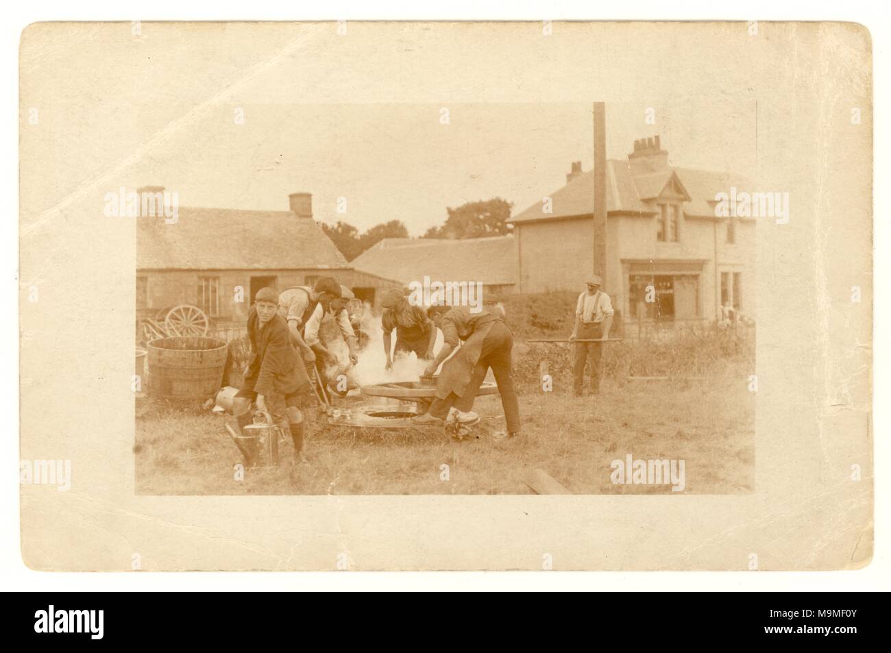 Original early 1900's postcard of craftsmen - wheelwrights /blacksmiths and apprentice making a cartwheel, fixing heated expanded iron tyres and - shrinking them onto the wheel with water,  outside the workshop on the village green. U.K. Circa 1920 Stock Photo