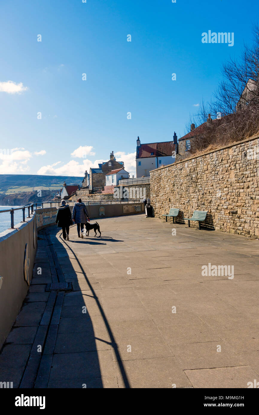 A couple with a dog ealking along top of the sea wall at Robin Hoods Bay with seats to enjoy the view and mosaics telling the local history Stock Photo