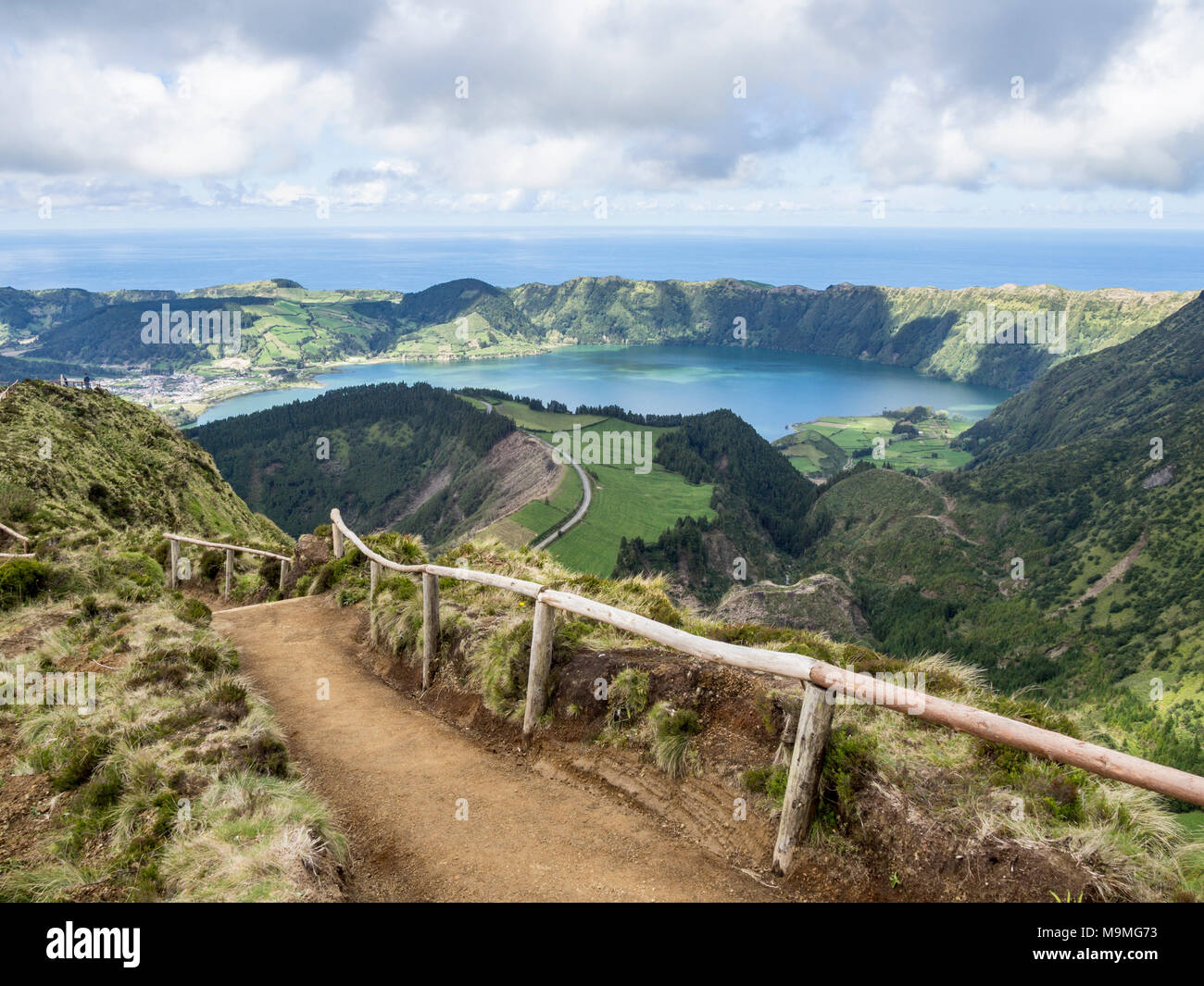 Path to the Road Below: A dirt path with wooden handrails winds over the hills to a spectacular viewpoint high over Sete Cidades and seems to connect to a road far below. Stock Photo
