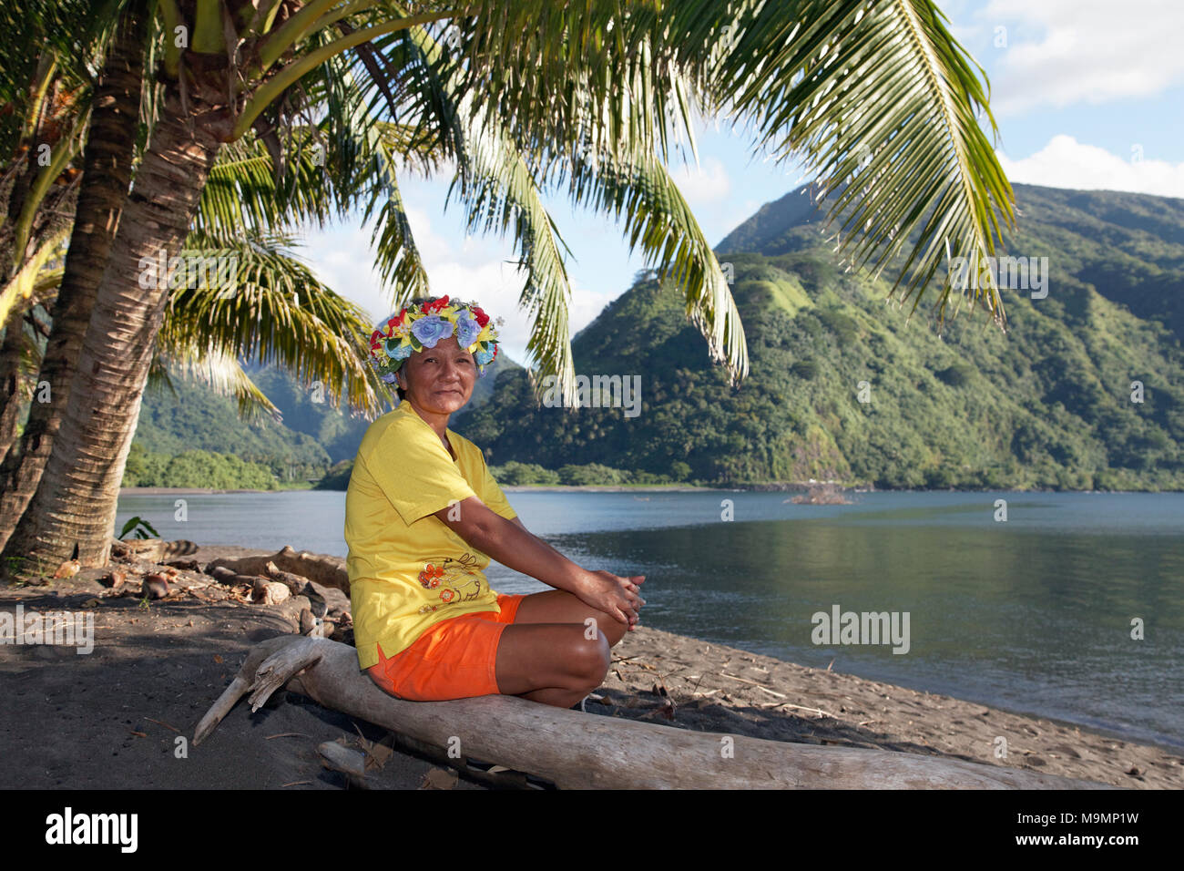 Old woman with wreath of flowers sitting under palm trees on tree trunk, coast near Taurita, Tahiti Iti, society islands Stock Photo