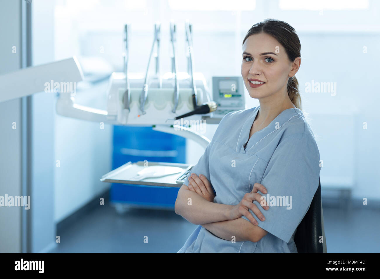 Nice workplace. Charming young female dentist posing in her office and smiling at the camera while folding her arms across the chest Stock Photo