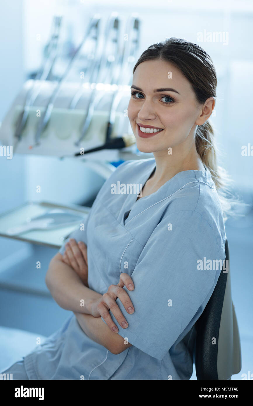 Love my workplace. Pretty young female dentist posing in front of the dental machine in her office and folding her arms across her chest while smiling Stock Photo