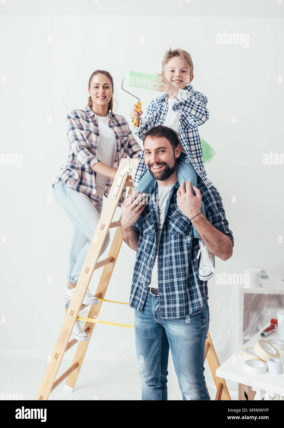 Happy family renovating their new home, they are posing together with a ladder and a paint roller, the father is piggybacking his son Stock Photo