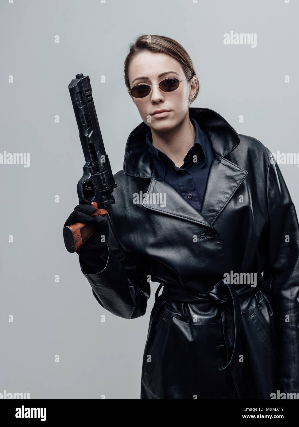 Cool female spy agent in black leather coat, she is holding a gun and posing Stock Photo