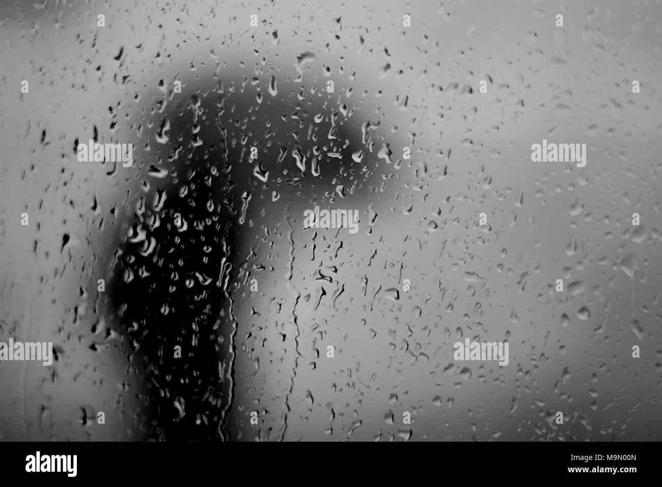 Person dressed all in black waliking hunched over under an umbrella seen through a rain covered window Stock Photo