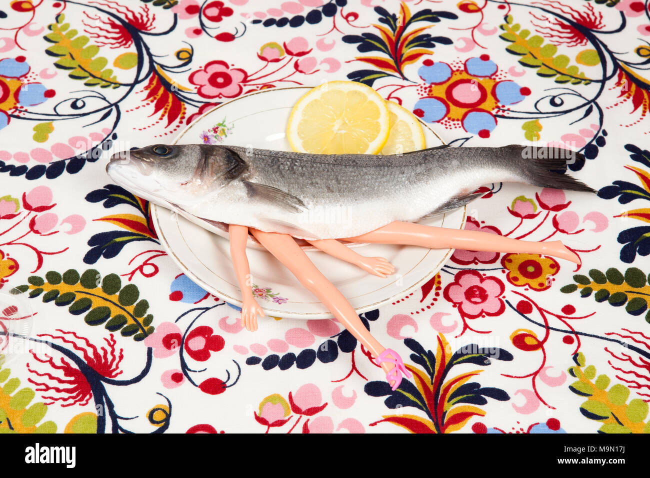 A bass fish with arms and legs of a doll inside on a flower plate.  Cannibalism and anthropomorphism on a floral fabric patterned background.  Quirky mi Stock Photo - Alamy