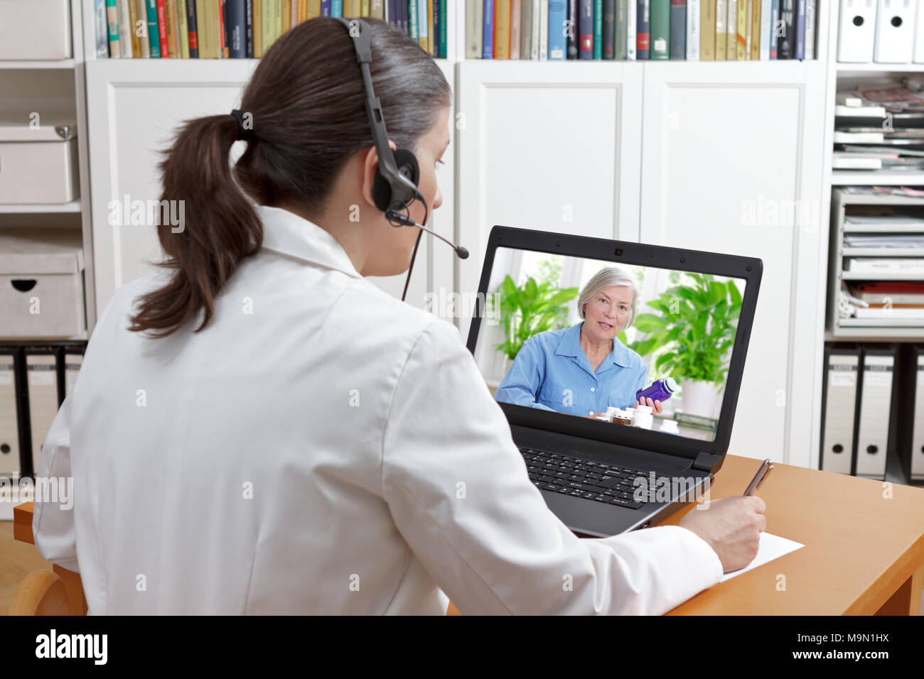 Doctor of geriatrics in her surgery office with headset in front of her laptop during a video call with a senior patient about her prescribed drugs. Stock Photo