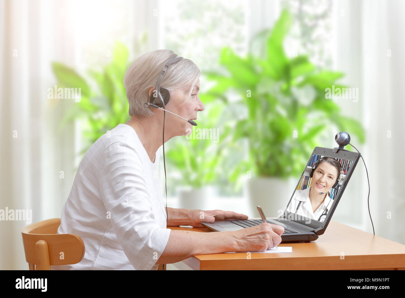 Senior woman in her living room in front of a laptop making notes during a video call with her female doctor Stock Photo
