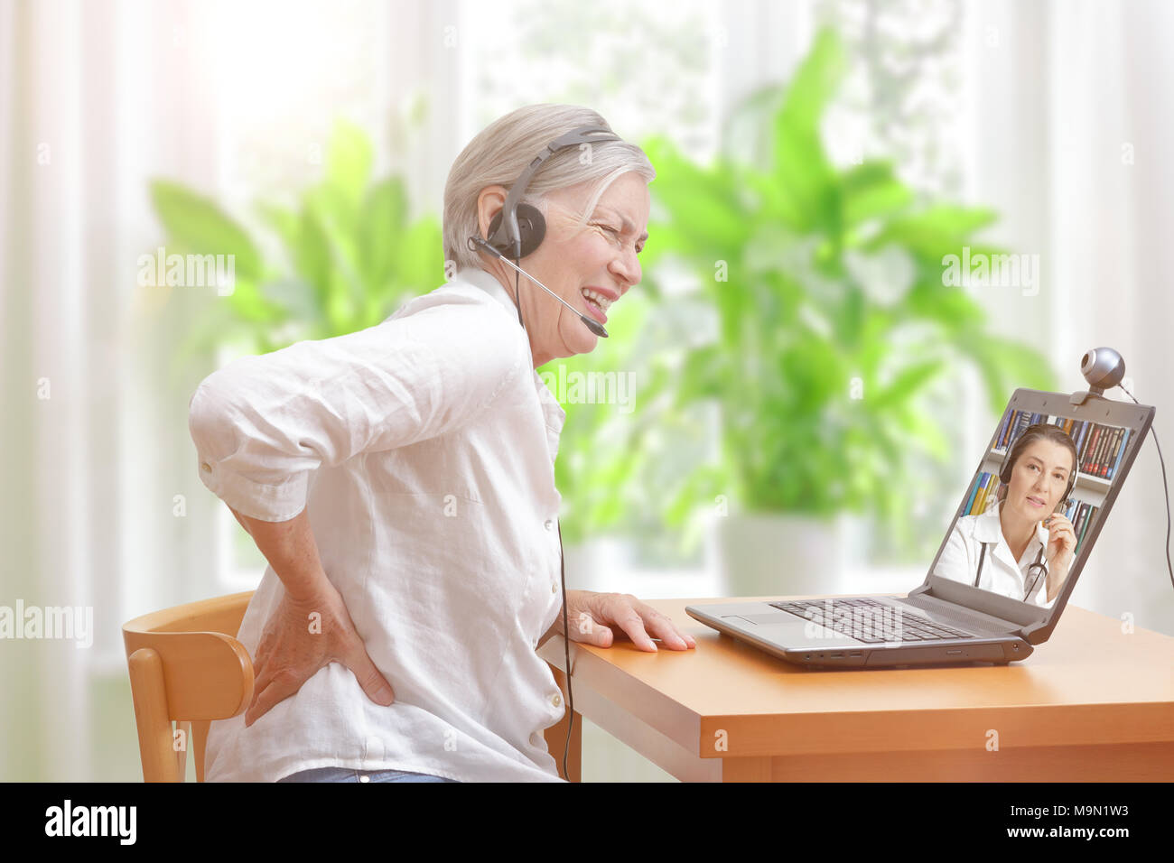 Senior woman with low back pain in her living room in front of a laptop making notes during a video call with her female doctor Stock Photo