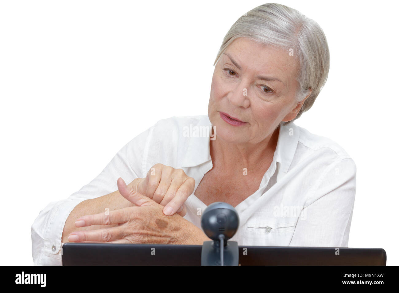 Elderly woman in front of a computer monitor with an attached webcam showing her doctor a mole on her hand during video call, isolated on white. Stock Photo