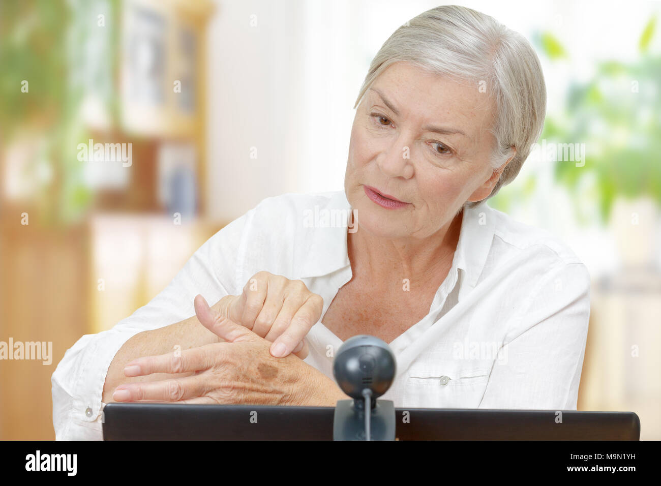 Senior woman in front of a computer monitor with an attached camera showing her doctor a mole on her hand during video call, telemedicine concept Stock Photo