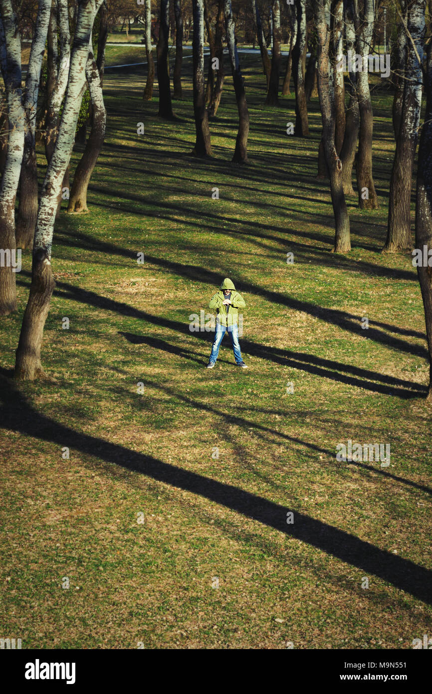 One mystery man in blue jeans and green hoodie standing surrounded with tall trees and it's shadows Stock Photo