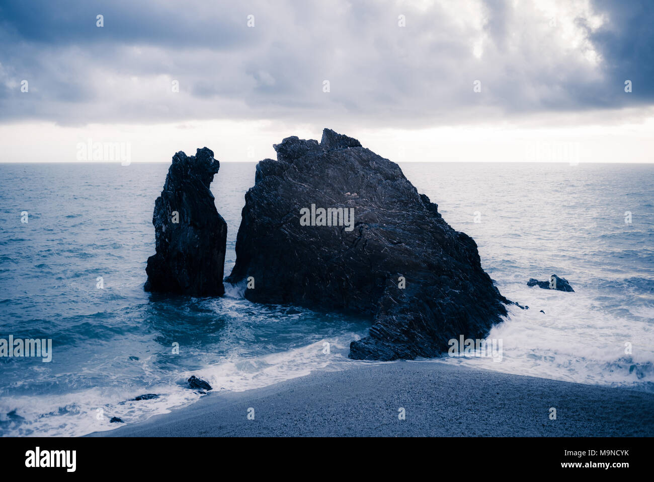 Black dark rock eroded cliff rising from the sea close to a sandy beach with dramatic cold light in a cloudy day Stock Photo