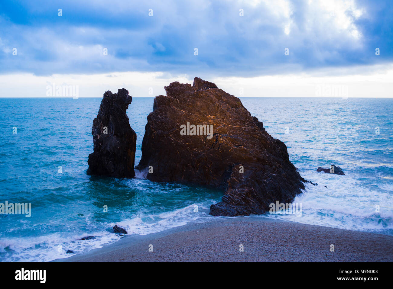 Black dark rock eroded cliff rising from the sea close to a sandy beach Stock Photo