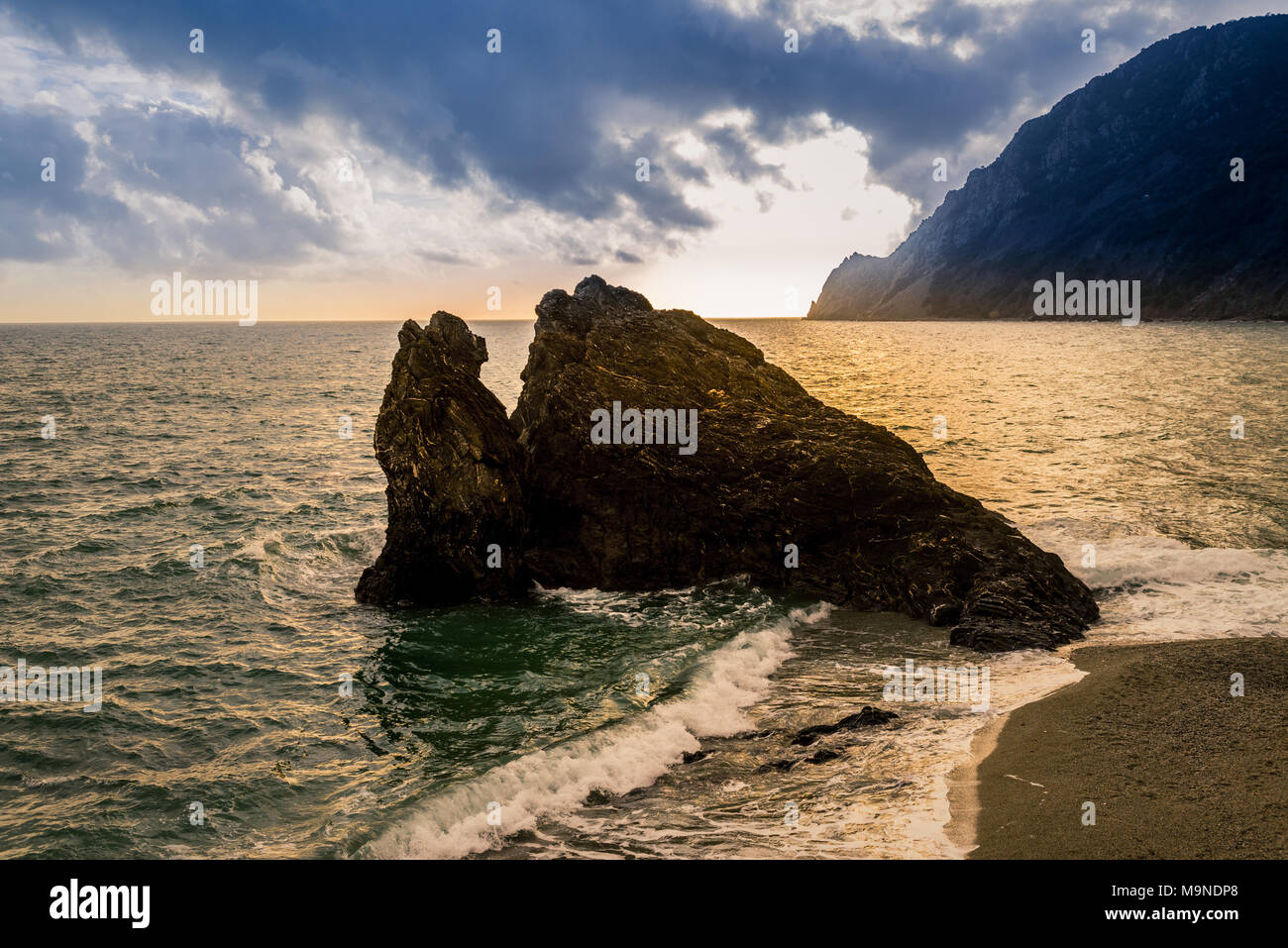Black dark rock eroded cliff rising from the sea ocean close to a sandy beach with mountain in the background Stock Photo