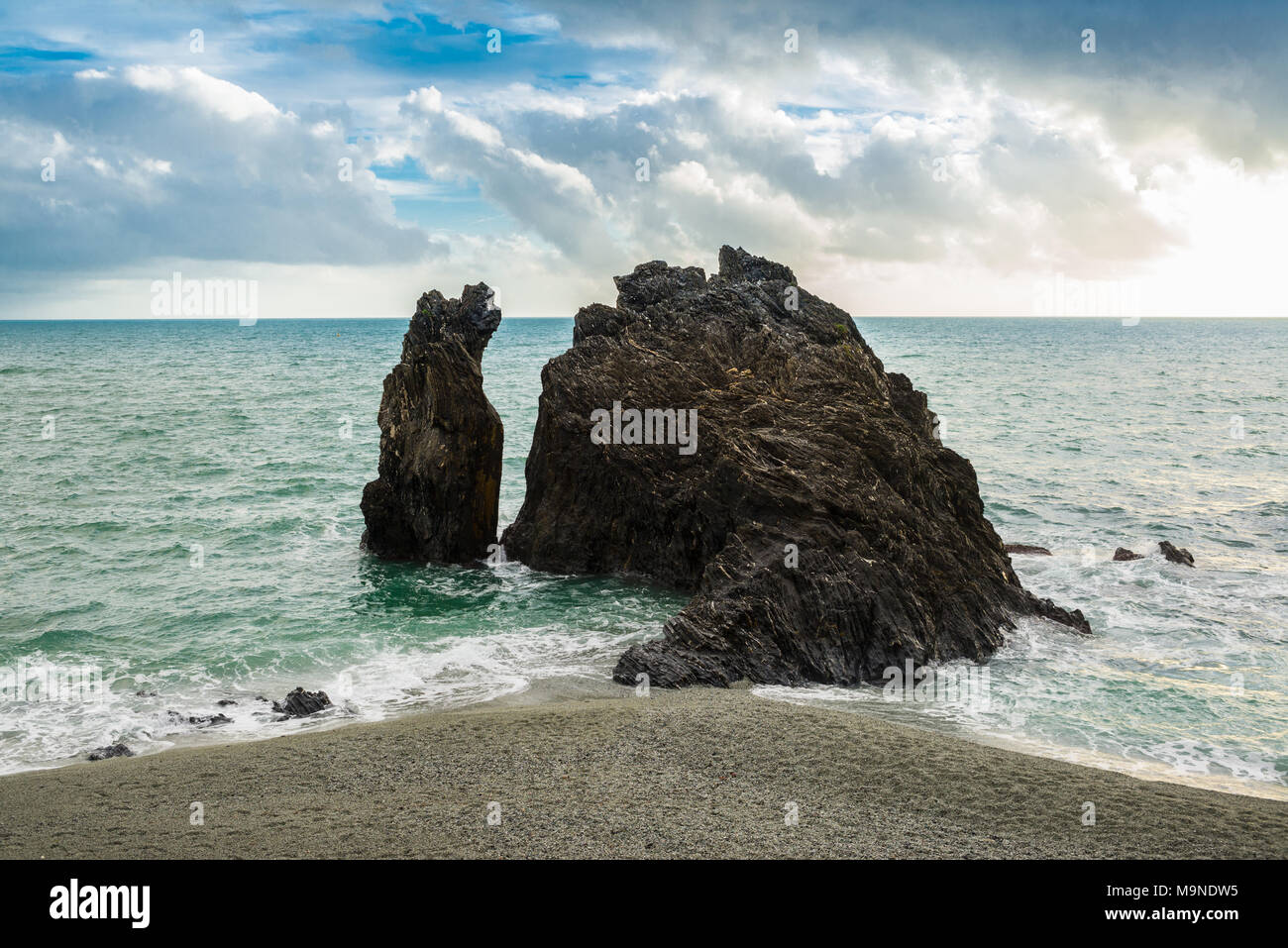 Black dark rock eroded cliff rising from the sea close to a sandy beach Stock Photo