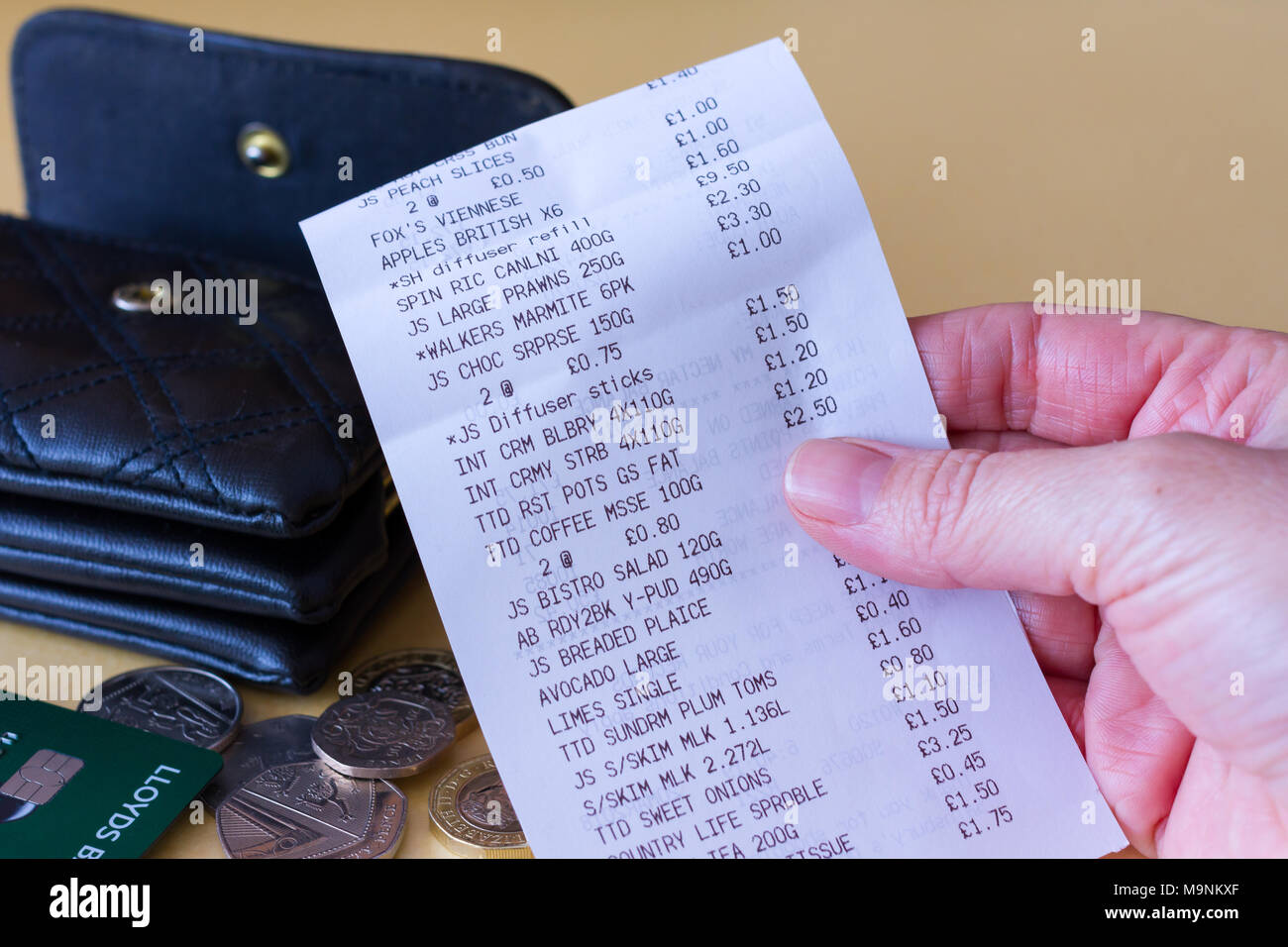 Female hand holding a supermarket grocery shopping till receipt with a purse, some coins & bank card in the background, United Kingdom Stock Photo