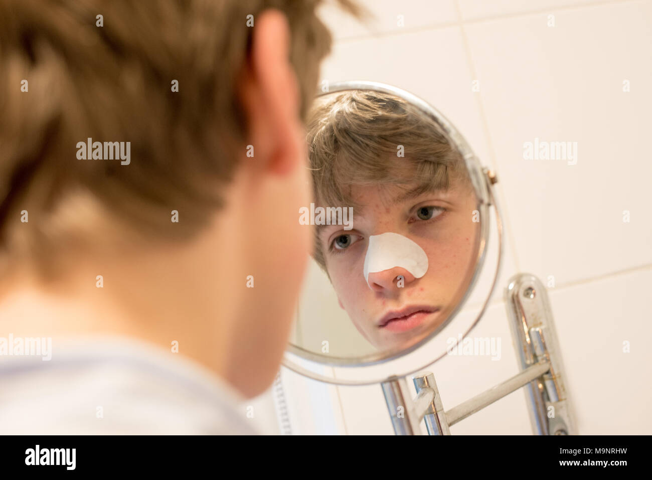 Fair-haired teenage boy looks in a bathroom mirror at his spots wearing a blackhead strip on his nose as part of a skincare routine Stock Photo