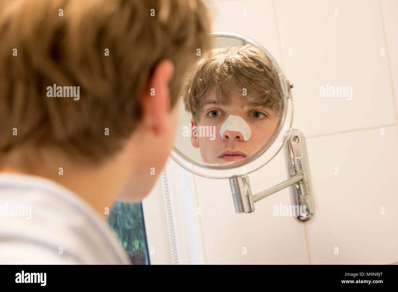 Fair-haired teenage boy looks in a bathroom mirror at his spots wearing a blackhead strip on his nose as part of a skincare routine Stock Photo