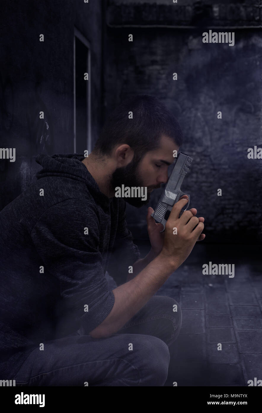 Young bearded man in a dark alley holds his head against a silver pistol Stock Photo