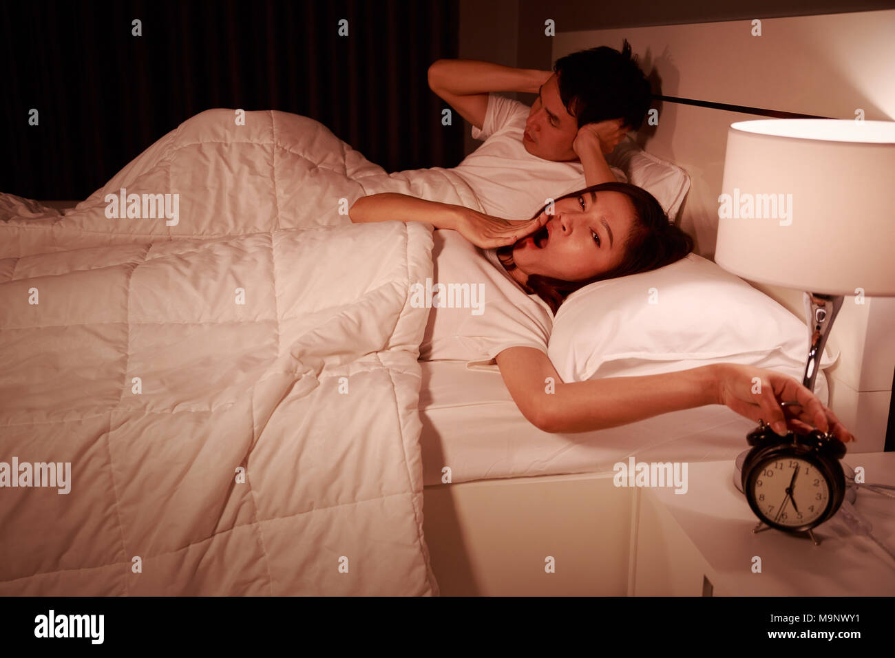 unhappy couple being awakened by an alarm clock in bedroom in the morning Stock Photo