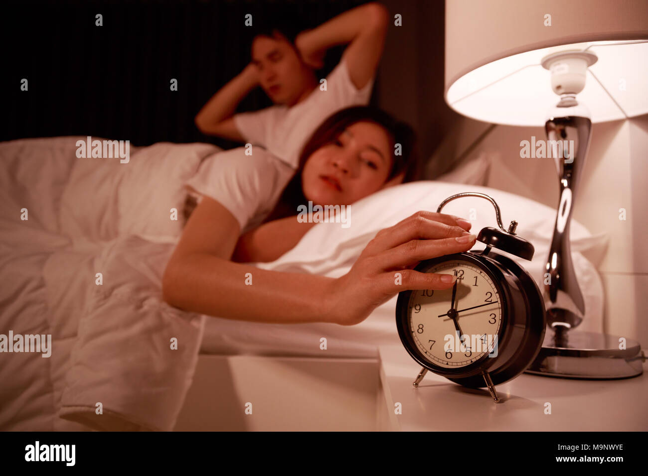 sleepy young couple in bed with extending hand to alarm clock in the morning Stock Photo