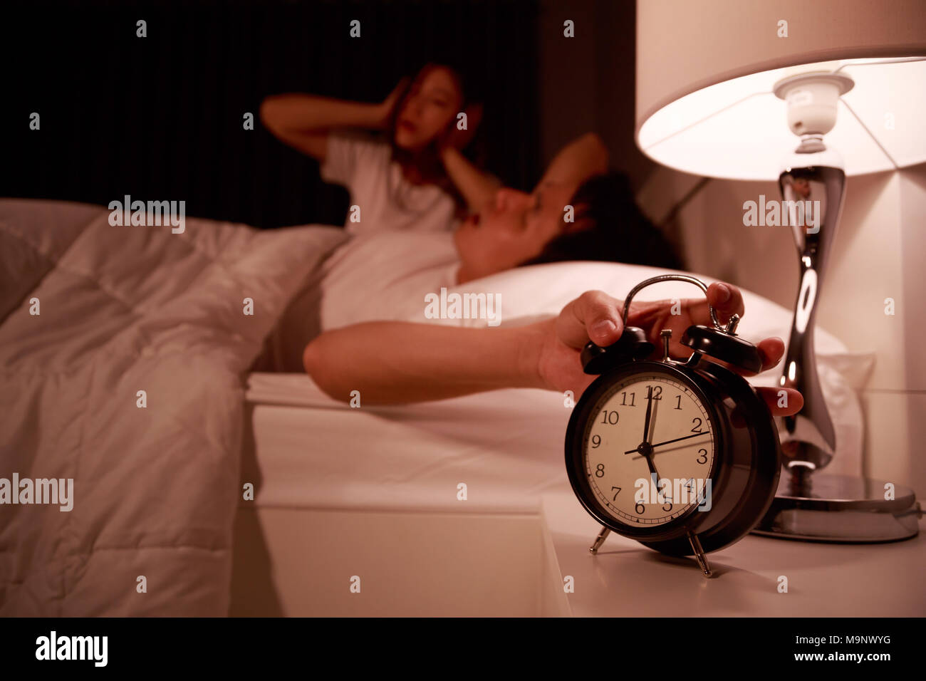 sleepy young couple in bed with extending hand to alarm clock in the morning Stock Photo